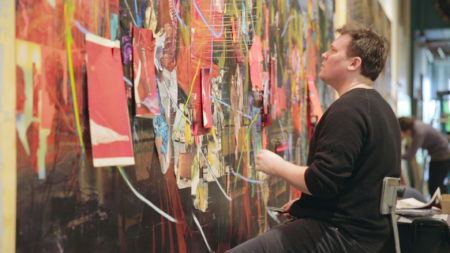 Elliot Hundley sitting on a chair in front of a large-scale piece hanging on the wall.