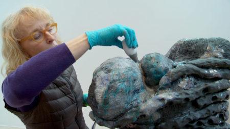 Arlene Shechet wearing blue gloves holding a squeeze bottle above a sculpture.