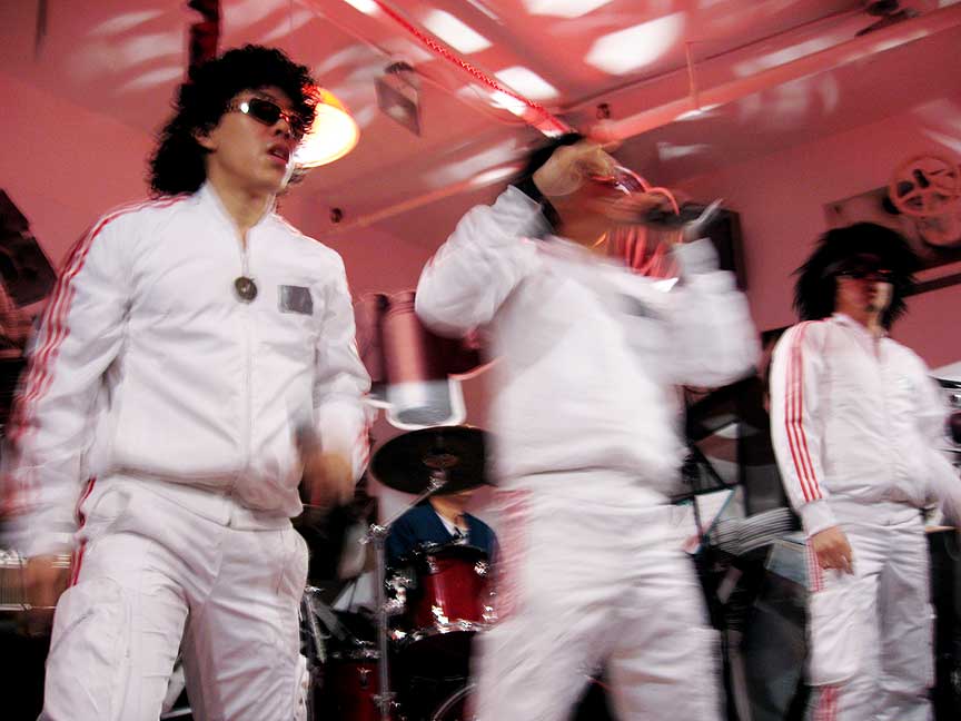 Three performers in white outfits, sunglasses, and shoulder length black wigs are captured mid-movement against a redlit background.