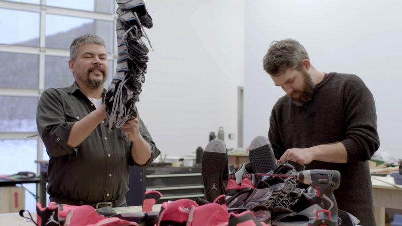 Brian Jungen and his assistant working in the studio on a piece constructed of sneakers, the piece Brian is holding is hanging from the ceiling.