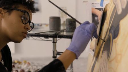 Jordan Casteel in her Brooklyn studio, wearing a blue latext glove and holding a paintbrush up to a canvas.