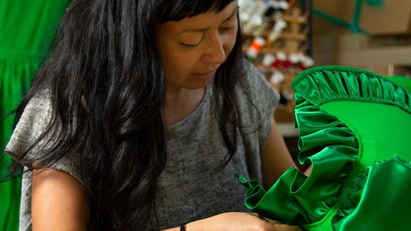 Stephanie Syjuco with long dark hair sews a shiny green ruffled fabric, with spools of thread visible in the background.