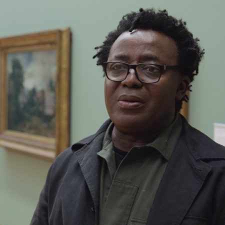 John Akomfrah wearing glasses, standing in a museum gallery with gold framed paintings in the background.