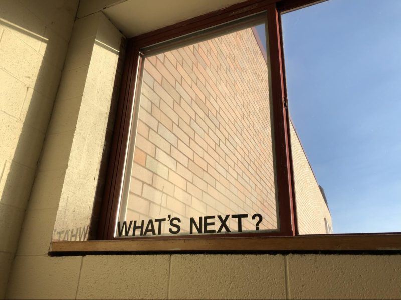 Photo of a school window from an interior viewpoint looking out to the brick exterior of the school building and a clear blue sky. Letter cutouts spelling the phrase "What's next?" are affixed to the window.
