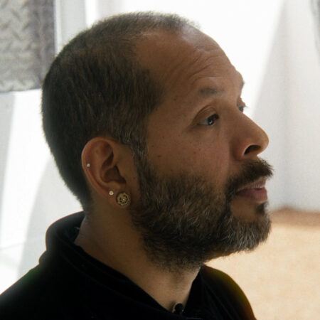 Guadalupe Maravilla with a beard and earrings along his ear looks intently at an object off-camera in a bright room.