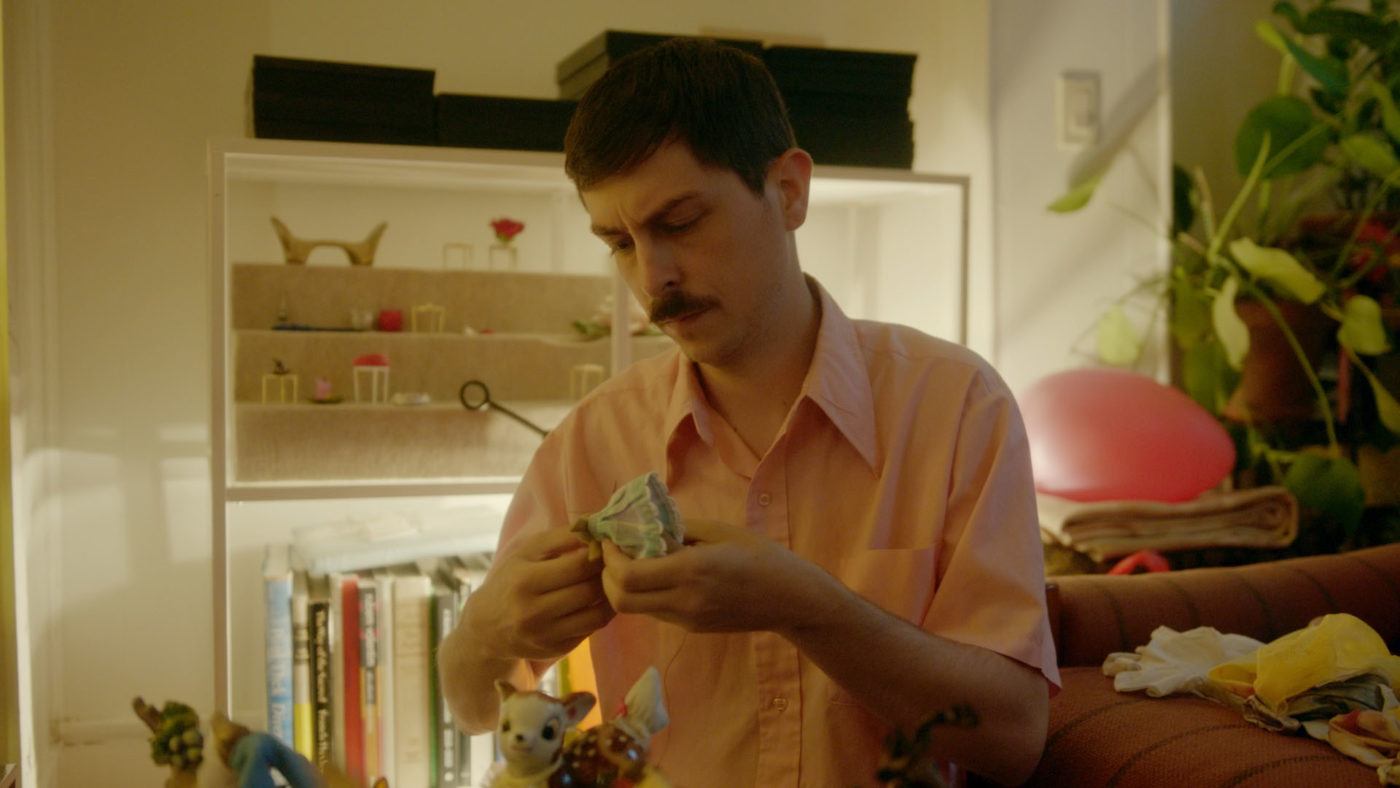 Adam Milner sitting in his home, holding a dress for one of his ceramic figurines.