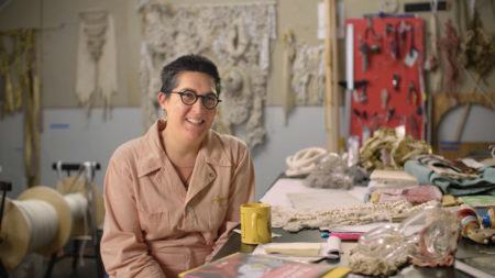 Tanya Aguiñiga sitting at a worktable in her studio. The worktable is covered in fabric and materials, behind her the walls are hung with weavings and tools.
