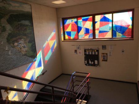 Colored tiles cover a hallway window an art installation by high school students. The reflection of the tiles drift down the wall of the staircase.