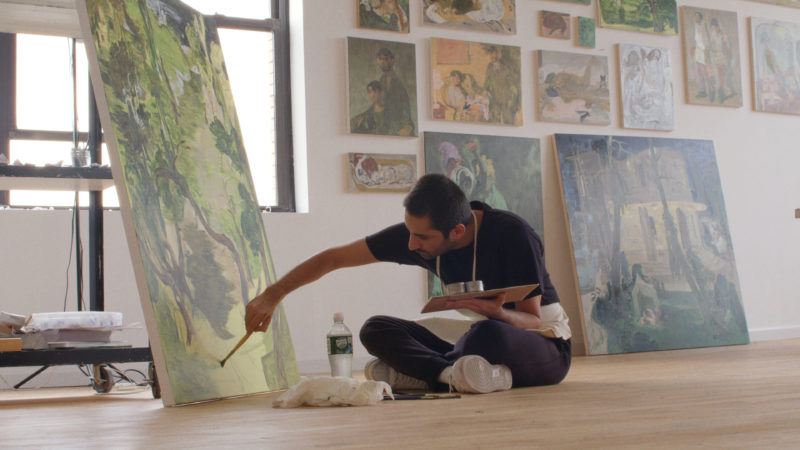 Salman Toor sitting cross-legged on the of floor of his studio working on a painting, one arm holding a palette and the other holding a paintbrush to the canvas.