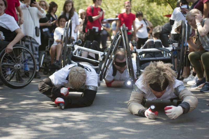 Participants of Pope.L’s "Conquest" crawling on the pavement, they were gloves, flashlihgts, and blindfolds with wheel chairs strapped to their backs. Crowds are on either side of them.