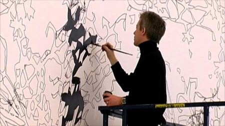 Matthew Ritchie wearing a black turtleneck painting black forms on a wall.