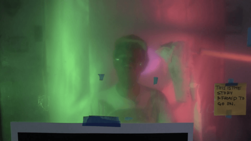 Still of Pope.L from a recent artist talk, the artist is reflected in a pink and green surface. A sticky note is in the bottom right that reads "This is the story afraid to go on."