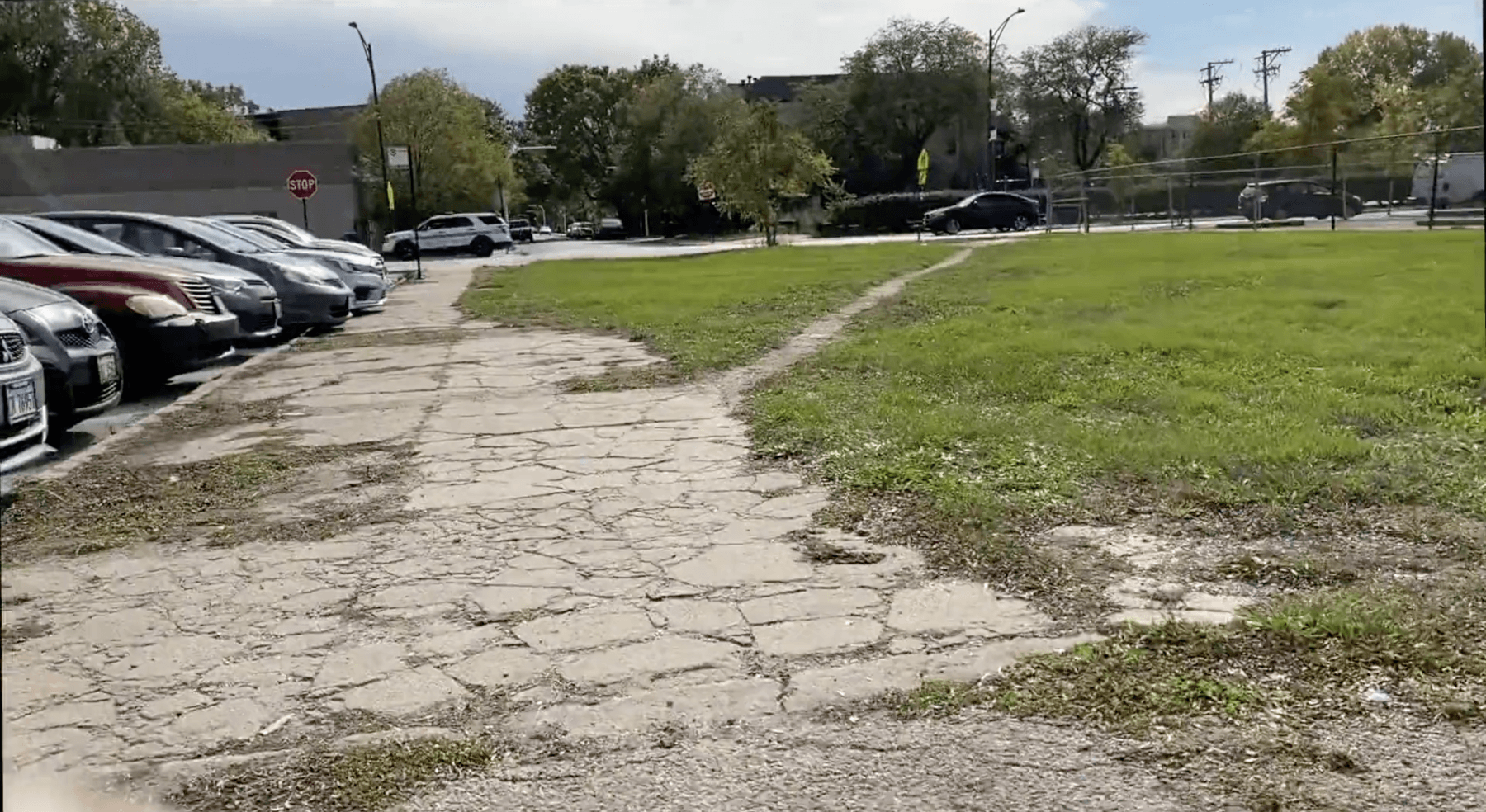 Image of a lot, on the right is grass with a small dirt pathway cut through it. On the right is broken up pavement with cars parked facing the curb.