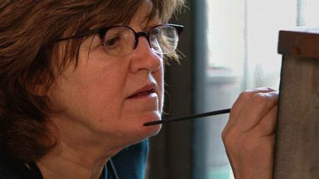 Closeup shot of Vija Celmins wearing glasses a holding a paintbrush up to a canvas.