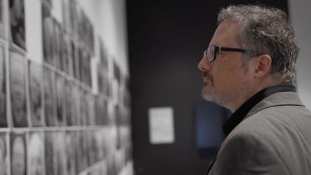 A profile shot of Rafael Lozano Hemmer wearing black glasses, and grey blazer, looking at a wall of black and white photos