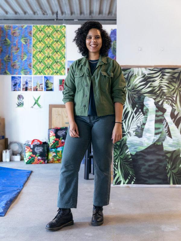 Joiri Minaya wearing a green overshirt, posing in her studio, with paintings hanging on the wall behind her.