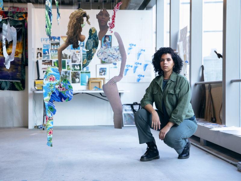 Joiri Minaya wearing a green overshirt, posing in her studio with knees bent and arms resting on knees.