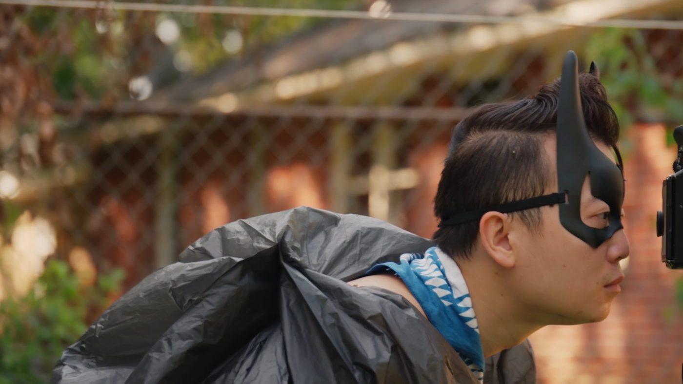 A profile shot of Tommy Kha wearing a batman mask and garbage bag over his clothes, looking through a camera.
