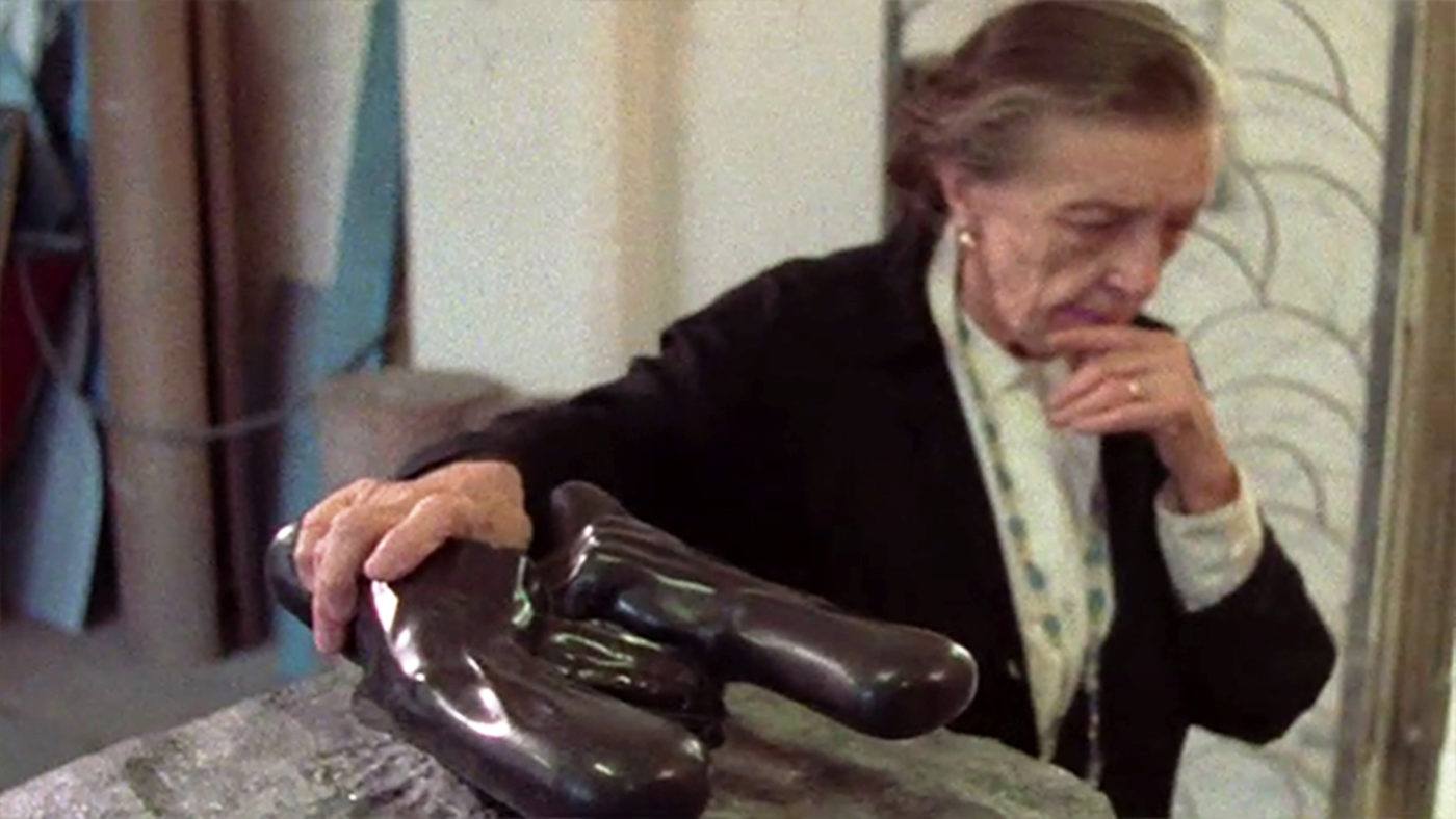Louise Bourgeois holding her hand to her chin looking down, while holding onto one of her sculptures sitting on a pedestal.