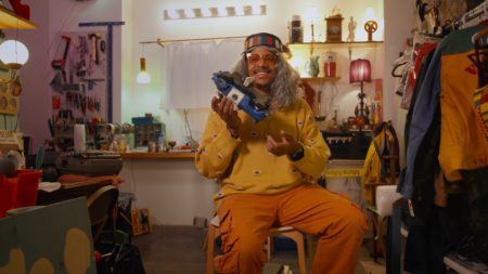 Azikiwe Mohammed wearing a grey wig and hat, yellow sweater and orange pants, and translucent yellow sunglasses holding a metal toy truck in his New Jersey studio. The studio in the background is filled with lots of different objects as well as tools hanging on a pegboard on the wall.