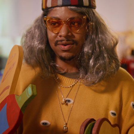 Closeup shot of Azikiwe Mohammed wearing a grey wig and hat, and translucent yellow cat-eye sunglasses.