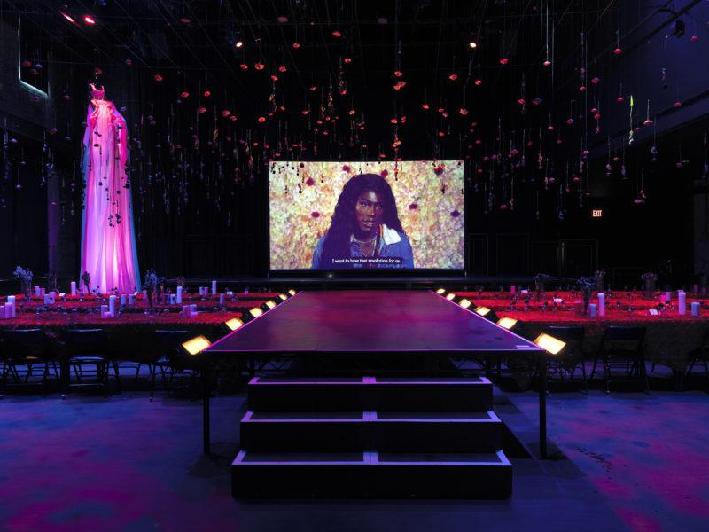 A runway extended into the picture, ending in a stage and a projection screen, upon which Lexii Fox is projected, mid speech.