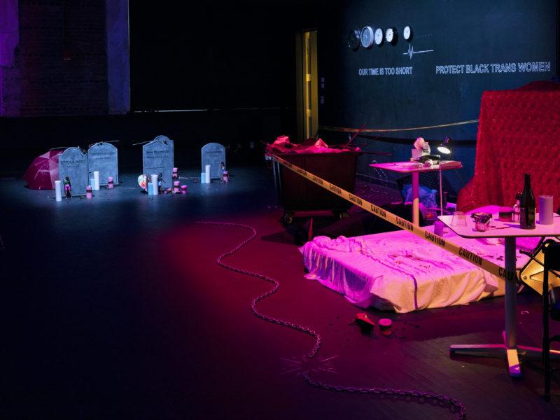 Multiple installations by Lexii Foxx at Performance Space. A bed sits on the floor to the right with bedside tables on either side, table tops covered in assorted items, the installation is surrounded by police caution tape. In back, five tombstones are visible, surrounded by votive candles and flowers.