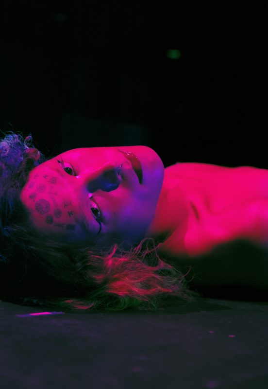 Jade Guanaro Kuriki Olivo is lying down nude, with a purple-red light cast on her, we look at her from the top of her head. She tilts her head backward to look at the camera.