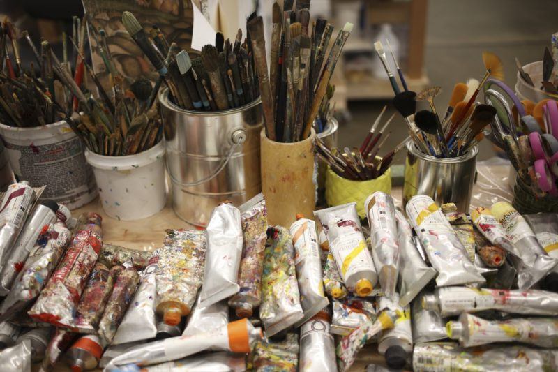 Table covered in cans filled with brushes and piled up tubes of paint.