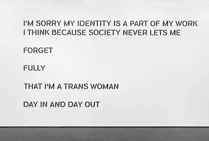 Installation view of a text piece, black text on a white wall reads, "I’M SORRY MY IDENTITY IS A PART OF MY WORK/ I THINK BECAUSE SOCIETY NEVER LETS ME/ FORGET/ FULLY/ THAT I’M A TRANS WOMAN/ DAY IN AND DAY OUT"