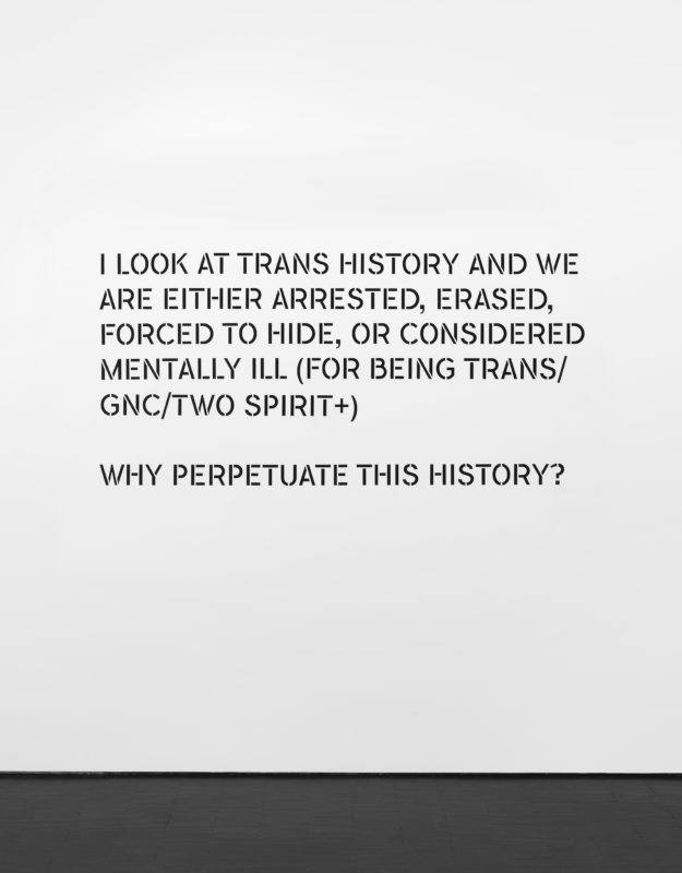 Installation view of a text piece, black text on a white wall reads, "I LOOK AT TRANS HISTORY AND WE ARE EITHER ARRESTED, ERASED, FORCED TO HIDE, OR CONSIDERED MENTALLY ILL (FOR BEING TRANS/GNC/TWO SPIRIT+) WHY PERPETUATE THIS HISTORY?"