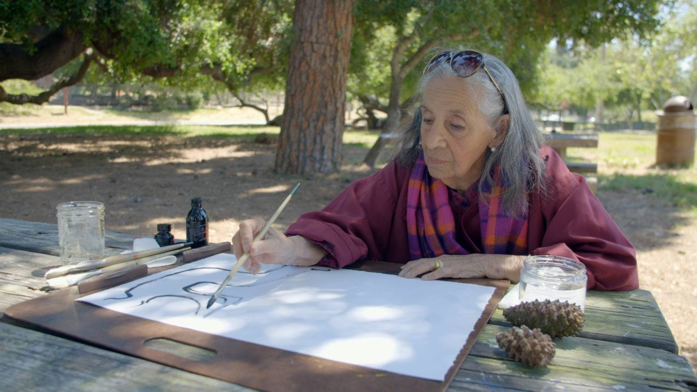 Luchita Hurtado sitting in a park at a picnic table painting on a white sheet of paper with black ink.