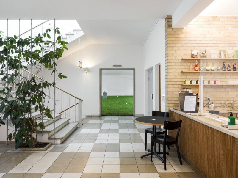 Through a wide doorway one sees fake grass and a tombstone in the distance. In the foreground, in front of the doorway, is a tiled floor, to the left of the doorway is a staircase going up and down, and to the right is a table with two chairs in front of a food and drink service bar.