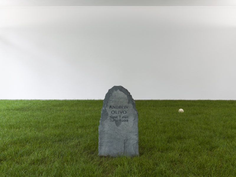 A tombstone sits in fake grass, with a ball sitting in the distance to the right of the tombstone. The tombstone reads "Andrew Olivo. June 7, 1989. June 7, 2018."
