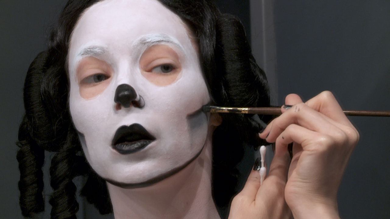 Mary Reid Kelley with her face painted white and nose and lips painted black, outlining her cheekbones in black with a paintbrush.