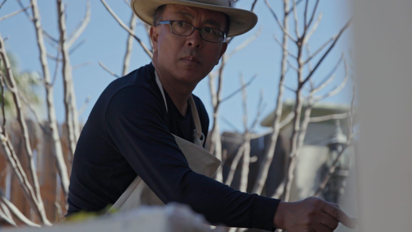 Liu Xiaodong wearing a brimmed hat and apron with trees in the background.
