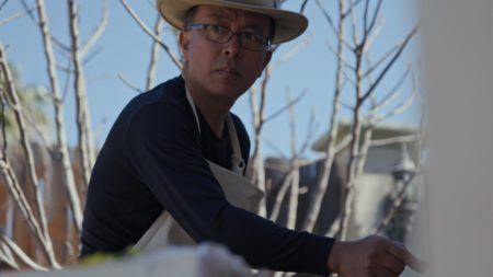 Liu Xiaodong wearing a brimmed hat and apron with trees in the background.