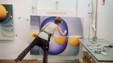 Loie Hollowell balanced on one foot leaning to work on a painting hung on a wall in her studio.