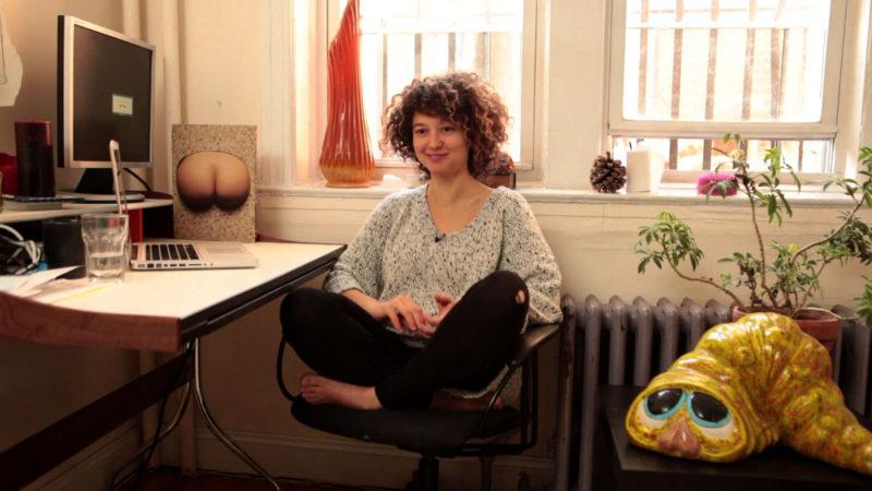 Mika Rottenberg sitting cross legged in a chair at her desk
