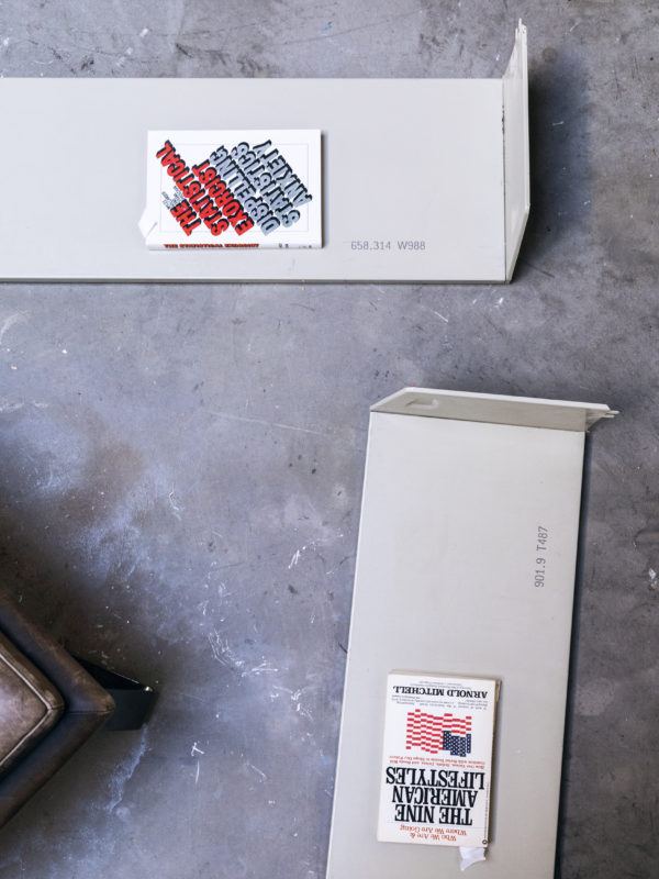 Two metal shelves sit on the floor of Rose's studio, perpendicular to one another, and atop each lays a book. The books are titled "The Statistical Exorcist" and "The Nine American Lifestyles."