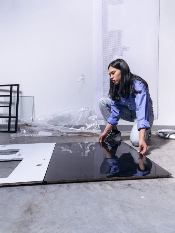 Artist Rose Salane arranges a sculptural work on the floor of her studio, behind her you can see a piece of paper hanging with a long list of words, another work.