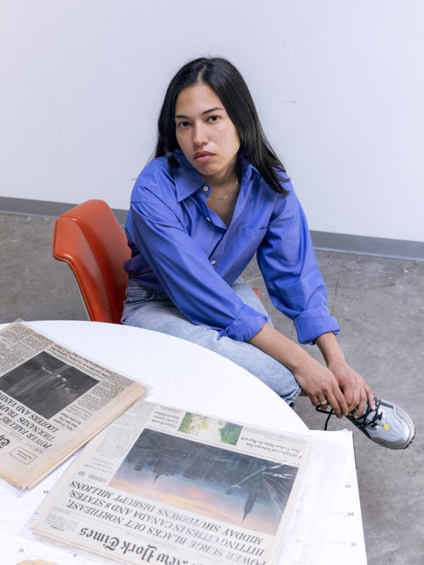 Artist Rose Salane sits legs crossed in an orange chair before a circular white table. Atop the table sit two newspapers, folded in half with their cover page facing up.