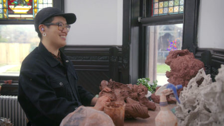Heid Lau wearing a black cap and black jacket in her studio at her Green-Wood Cemetery studio, surrounded by clay sculptures