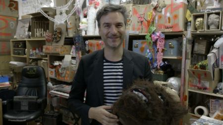 Marcel Dzama standing in his studio holding a werewolf costume head. The shelves behind him are full of boxes and supplies.