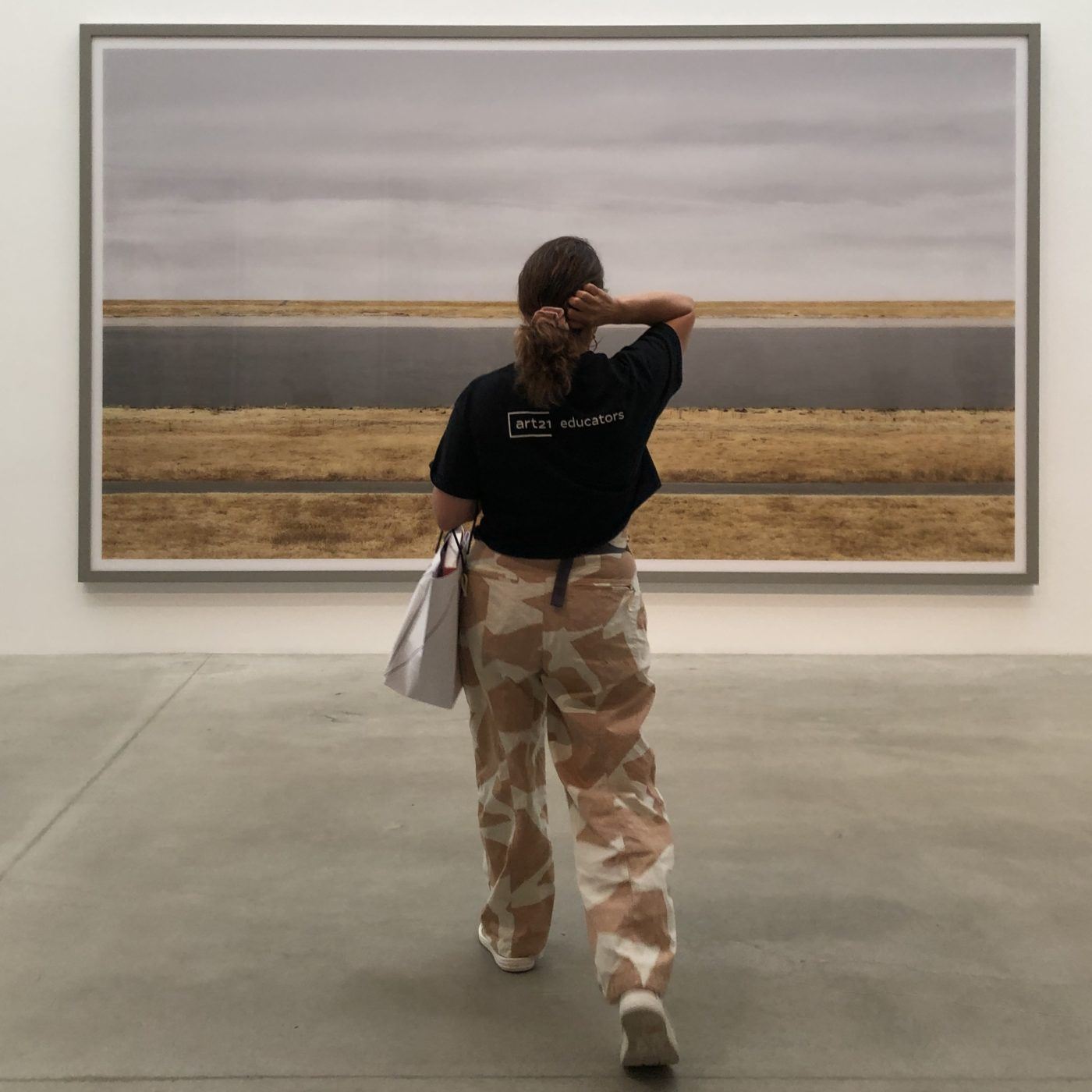 Woman with her arm raised to her head, wearing tan patterned pants and a black Art21 Educators t-shirt standing in front of a large-scale image of a road and gray sky.