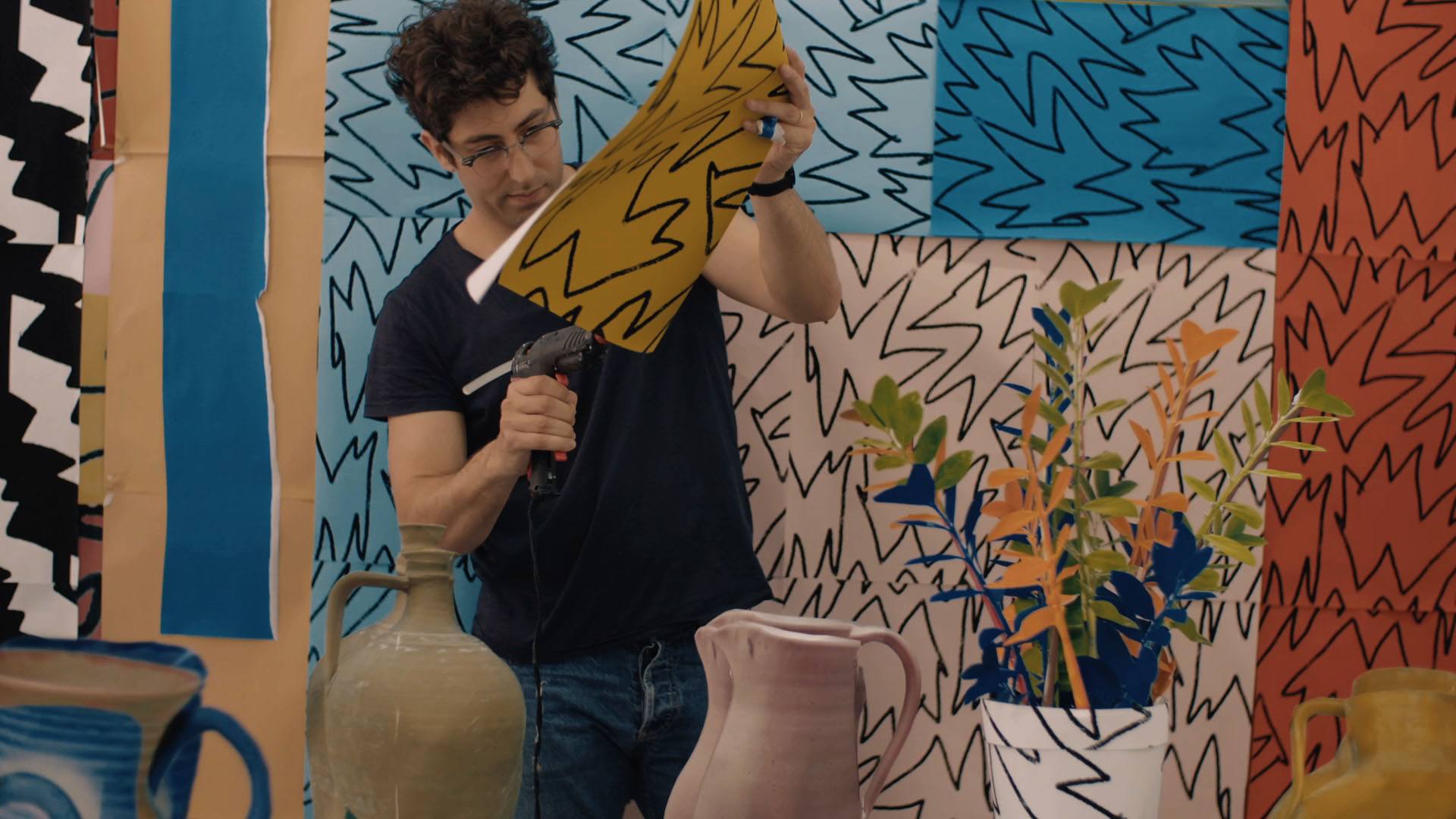 Daniel Gordon hot gluing a bright yellow paper with black outlined shapes drawn on it to a wall of multicolored papers with similar shapes drawn on them.