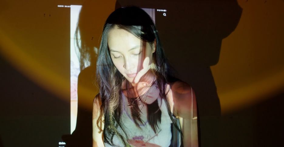Diane Severin Nguyen sitting in a dark room looking down with videos projecting over her face.