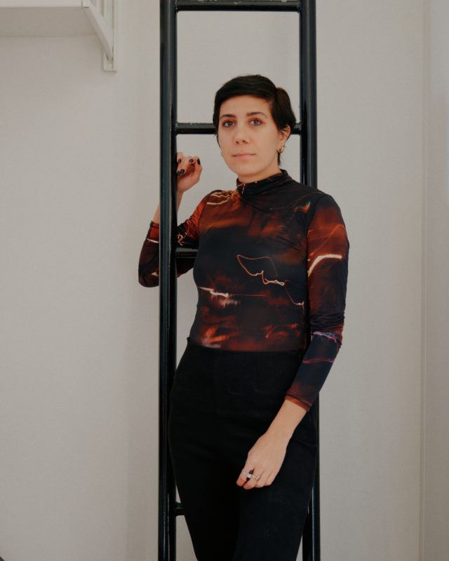 Artist Morehshin Allahyari stands in front of a ladder indoors wearing a red, orange, and gray long sleeved shirt with black pants.