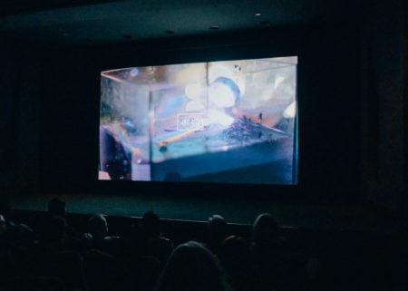 Audience seated in a theater with an abstracted image projected on the screen with the Art21 logo in the center.