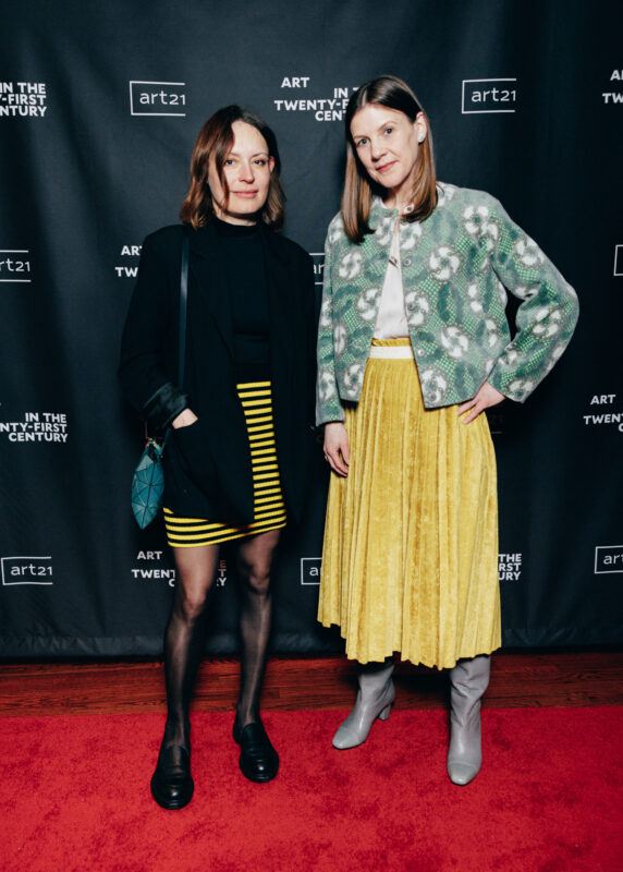 Paulina Pobocha and Maartje Oldenb posing for a photo in front of a Art21 backdrop. One in a black shirt yellow and black striped skirt black blazer and blue bag. other in white shirt blue and white blazer and yellow skirt.